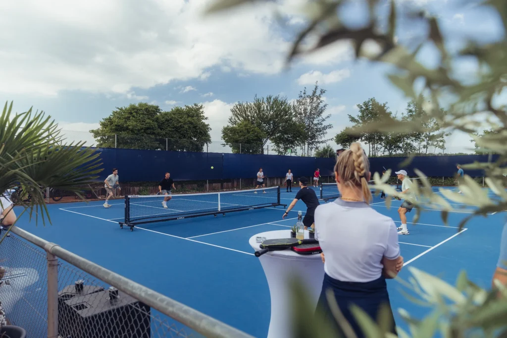 Blick von außen auf ein Pickleball-Turnier. Zu sehen sind drei Courts, auf denen Matches ausgetragen werden. Im Vordergrund stehen Zuschauer.